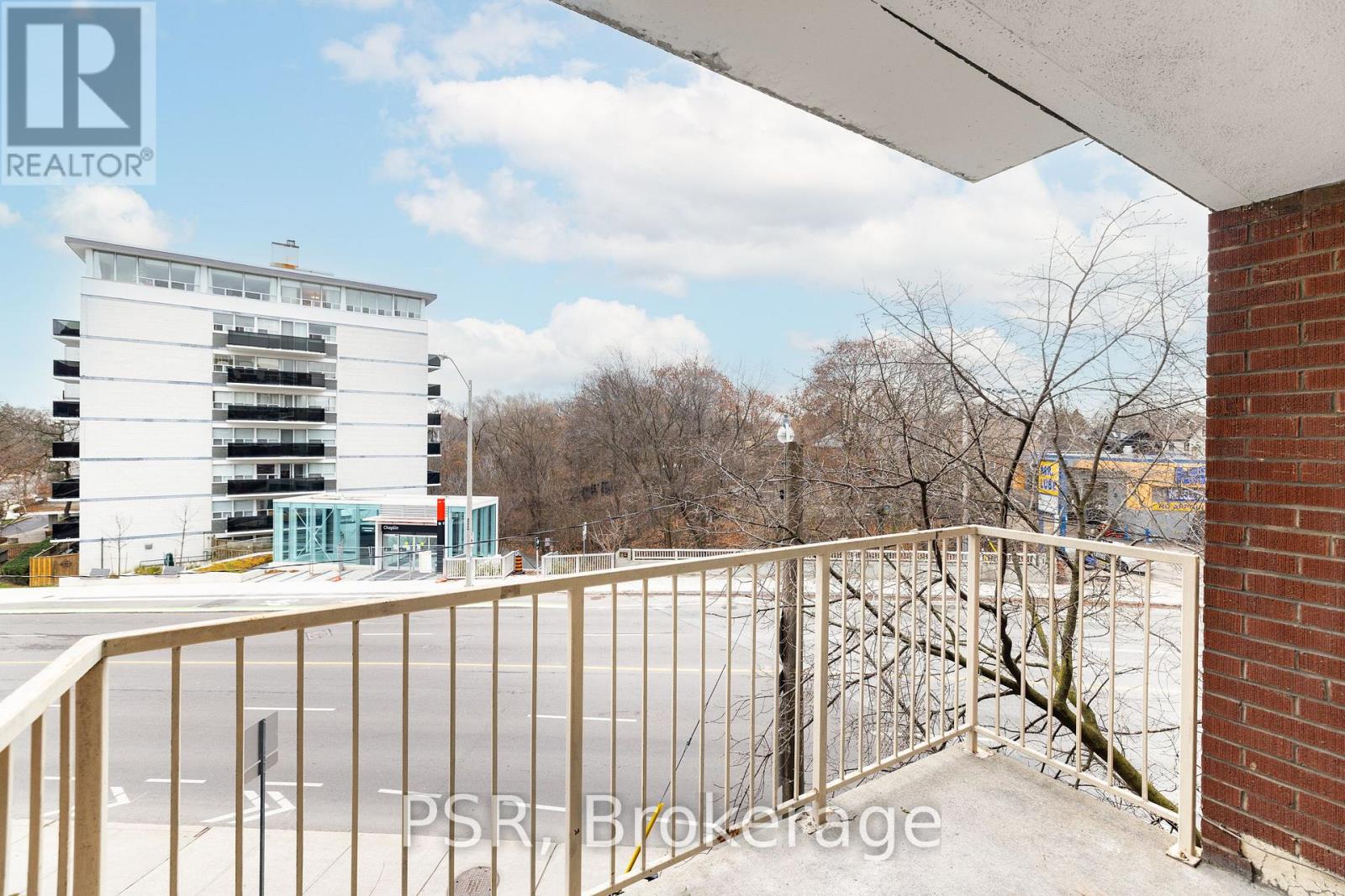 A balcony with a metal railing overlooks a parking lot and a multi-story building.