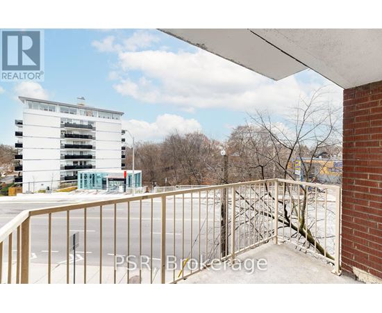 A balcony with a metal railing overlooks a parking lot and a multi-story building.
