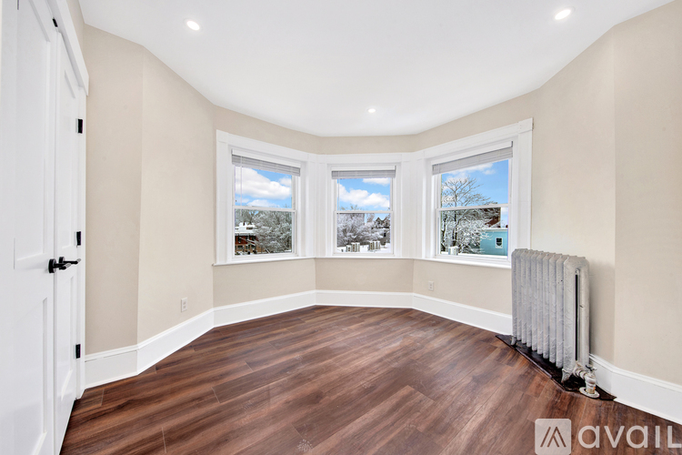 A room with a white door, a window with a view of the sky and trees, and a radiator on the floor.
