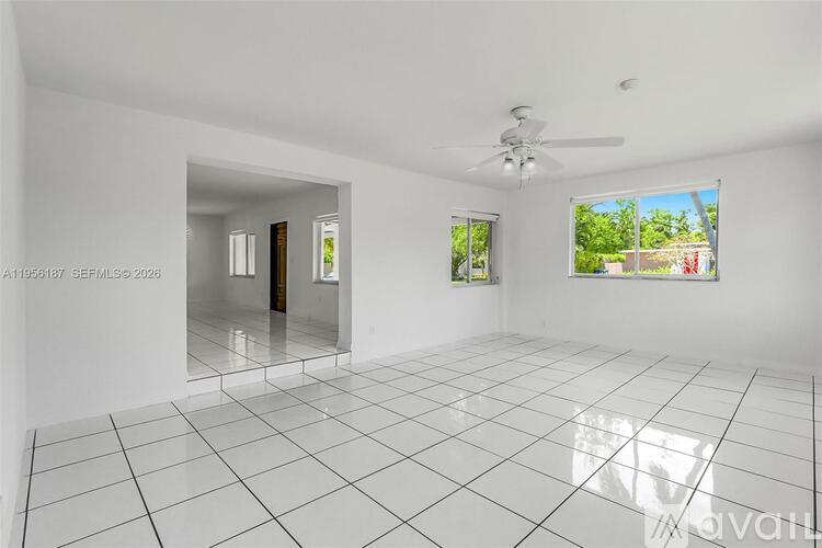 A spacious, empty room with white tiled floors and walls, a ceiling fan, and a view of greenery outside through a window.