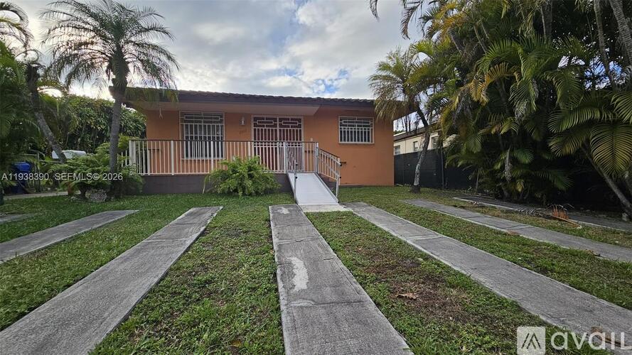 A house with a brown exterior and a white door is surrounded by a grassy area and palm trees.