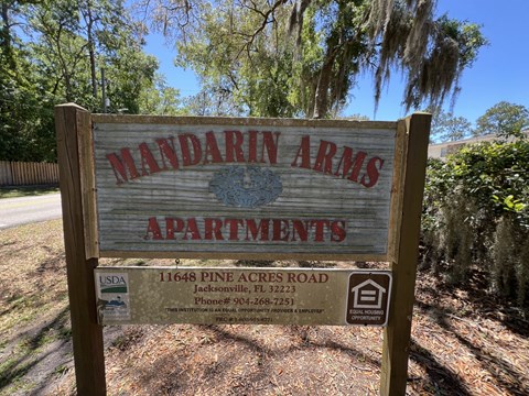 A sign for Mandarin Arms Apartments is displayed in front of a wooded area.