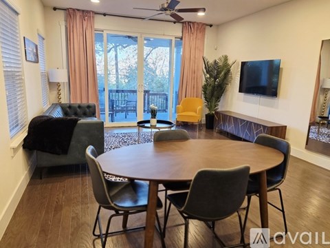 A living room with a wooden table and chairs, a television, and a balcony with a view of trees.