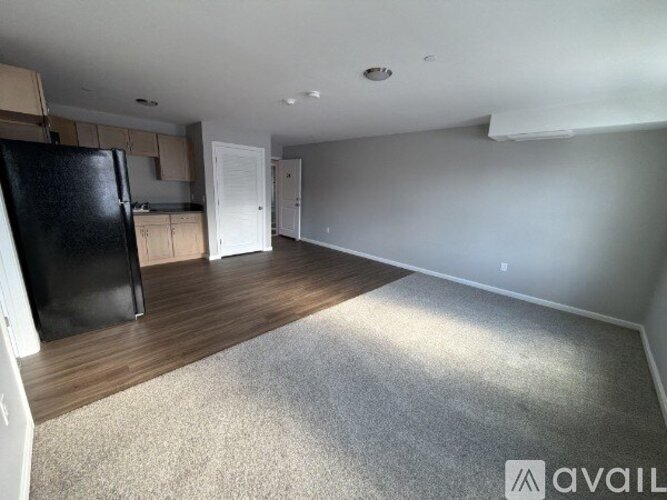 A spacious room with a black fridge, wooden floors, and white walls.