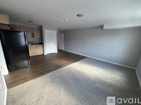 A spacious room with a black fridge, wooden floors, and white walls.