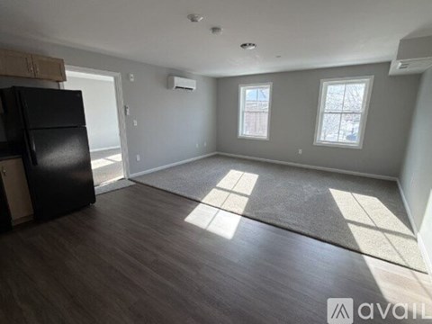 A spacious room with a black fridge, wooden floors, and two windows.