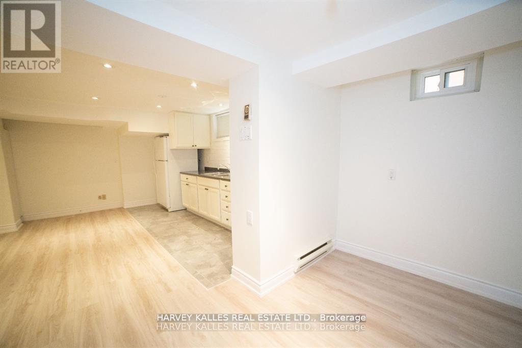 A spacious room with wooden flooring and white walls, featuring a kitchen area with cabinets and a window.