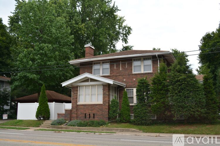 A house with a brown brick exterior and a white window is surrounded by greenery.