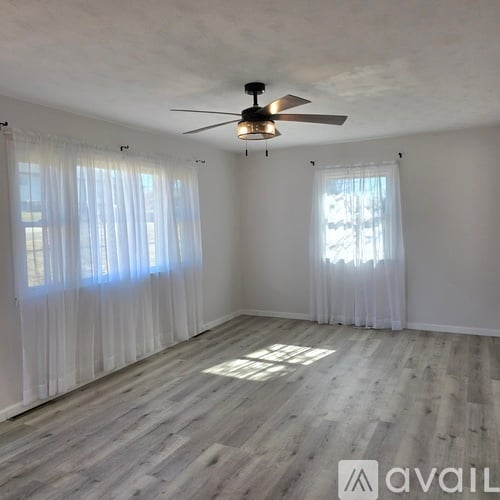 A room with a ceiling fan and light, two windows with curtains, and wooden flooring.
