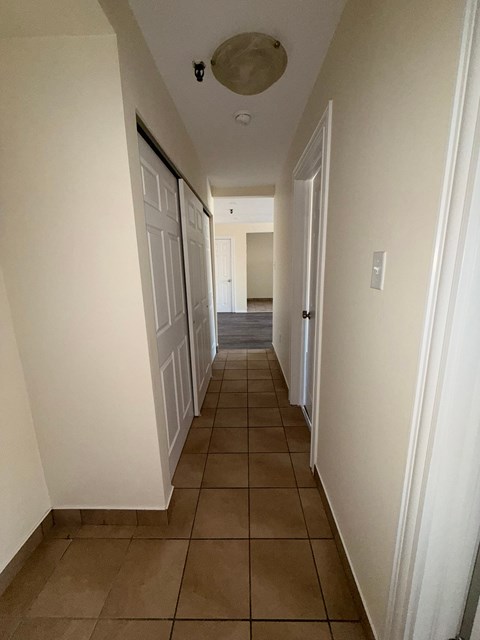 A long hallway with white walls and brown tile flooring.