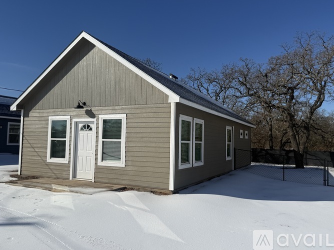A small house with a grey siding and a white door is surrounded by snow.
