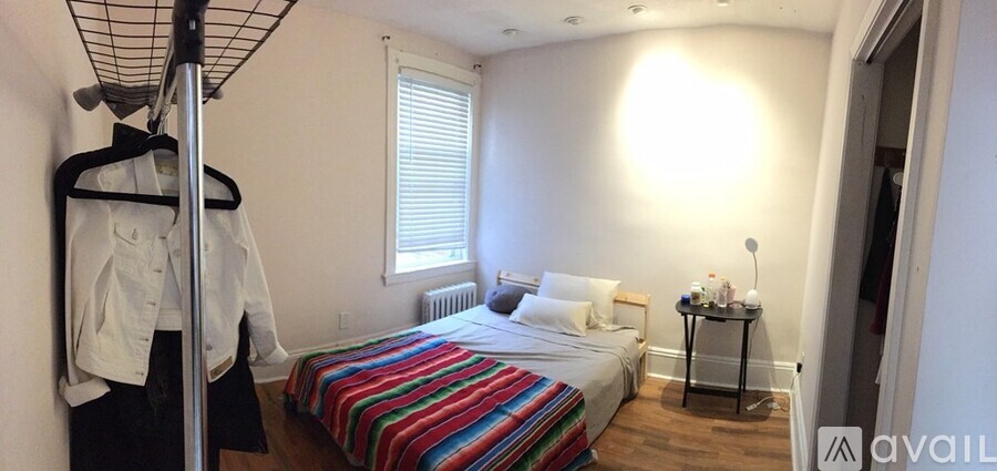 A bedroom with a bed, a window with blinds, a nightstand with a lamp, and a clothing rack.
