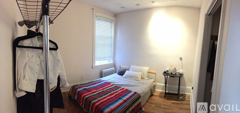 A bedroom with a bed, a window with blinds, a nightstand with a lamp, and a clothing rack.