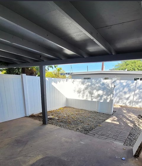 A concrete patio area under a roof with a white fence and a gravel area in the middle.