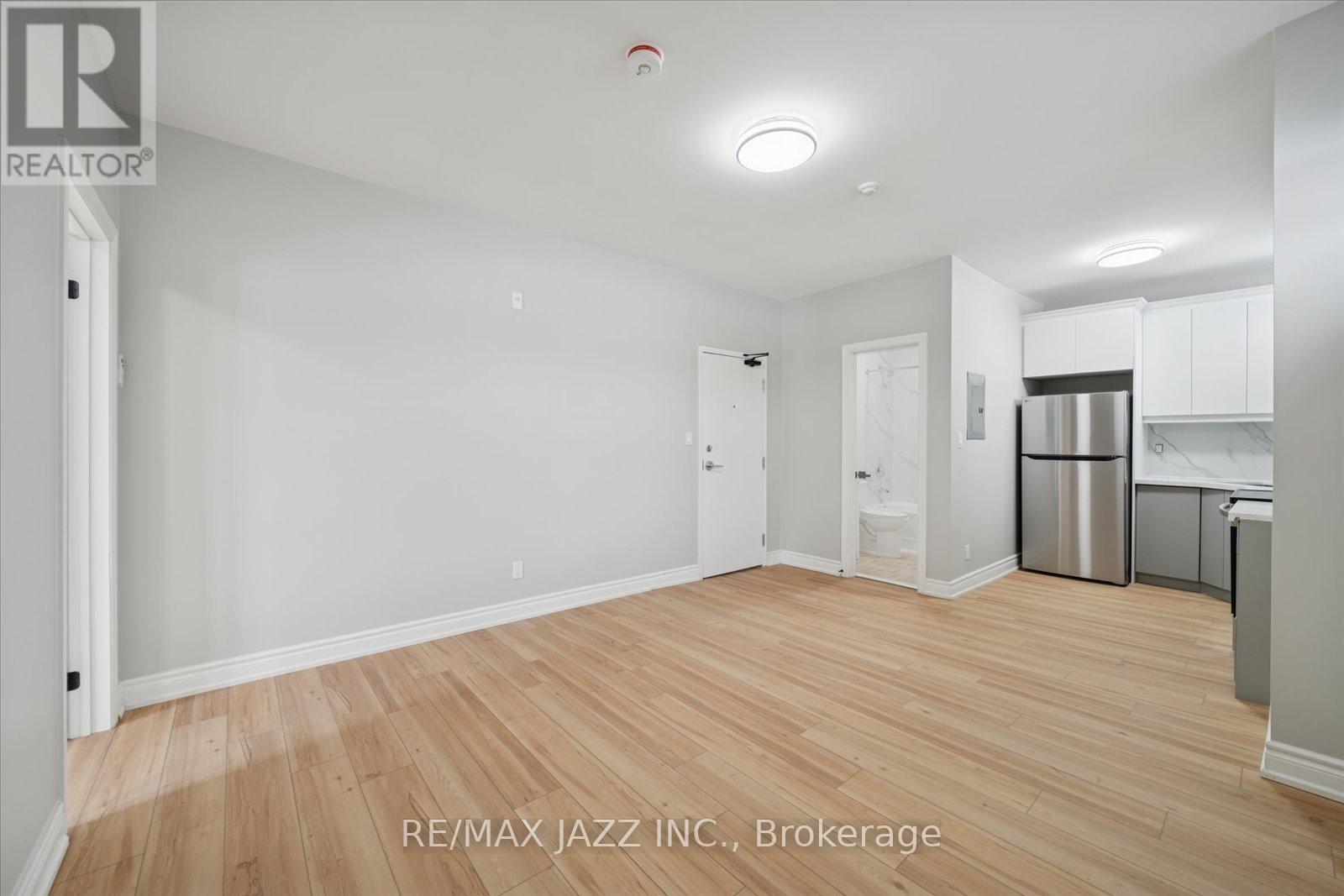 A spacious room with wooden flooring and white walls, with a kitchen area visible in the background.