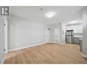 A spacious room with wooden flooring and white walls, with a kitchen area visible in the background.