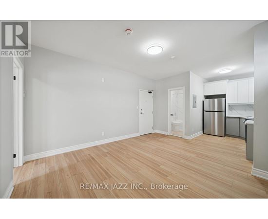 A spacious room with wooden flooring and white walls, with a kitchen area visible in the background.
