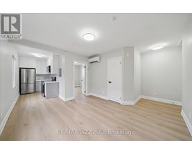 A spacious room with wooden flooring and white walls, featuring a kitchenette with stainless steel appliances.