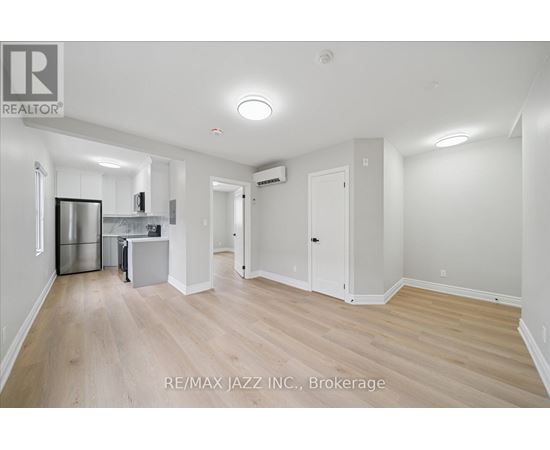 A spacious room with wooden flooring and white walls, featuring a kitchenette with stainless steel appliances.