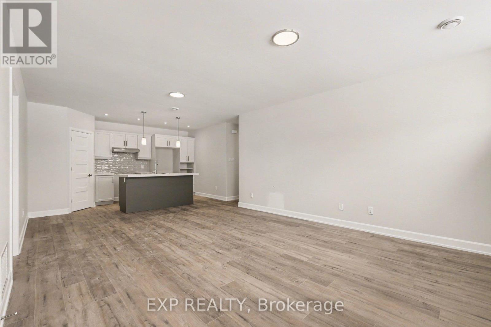 A room with a wooden floor and white walls, with a kitchen area visible in the background.