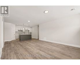 A room with a wooden floor and white walls, with a kitchen area visible in the background.