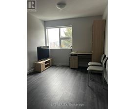 A room with a grey floor and a window with a view of trees and buildings outside.