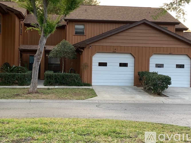 A brown house with two white garage doors and a tree in front.