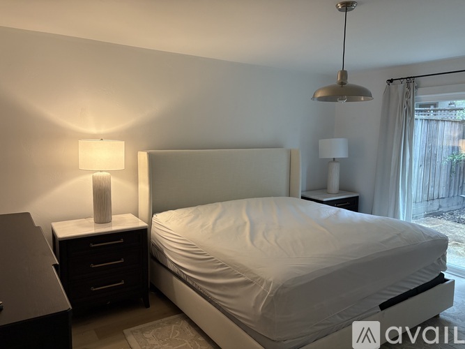 A bedroom with a white bed, a nightstand with a lamp, and a window with curtains.