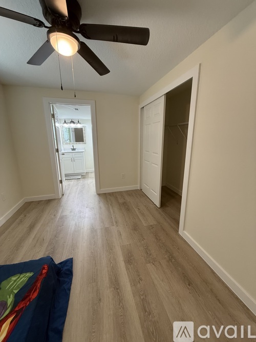 A room with a ceiling fan and light, wooden flooring, and a doorway leading to another room.