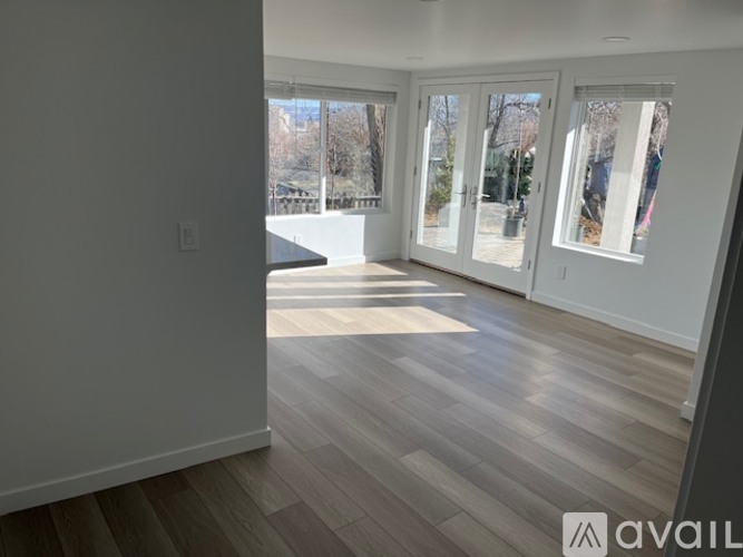 A room with wooden flooring and white walls, with a view of trees outside through the windows.