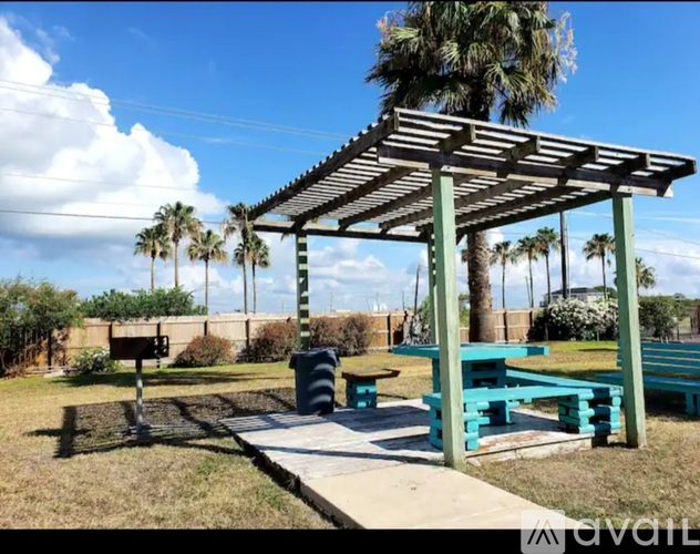 A wooden pergola with blue benches is in a grassy area with palm trees.