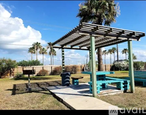 A wooden pergola with blue benches is in a grassy area with palm trees.