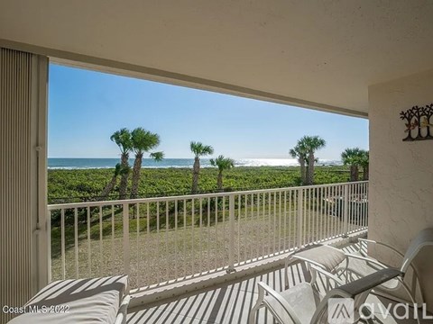 A balcony with white chairs overlooks a beach with palm trees.