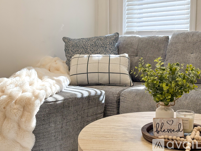 A grey couch with a checkered pillow and a white fluffy throw on the left side, a wooden table with a vase of green plant, a candle, and a sign that says "blessed" in the middle of