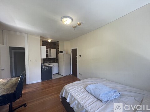 A bedroom with a bed, chair, and kitchen area in the background.