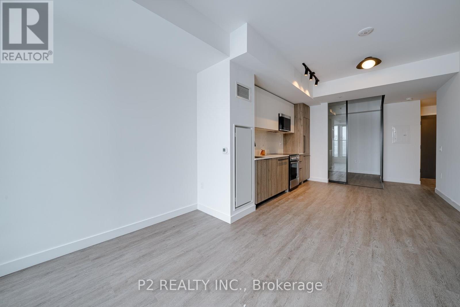 A spacious, empty room with wood flooring and white walls, with a kitchen area visible in the background.