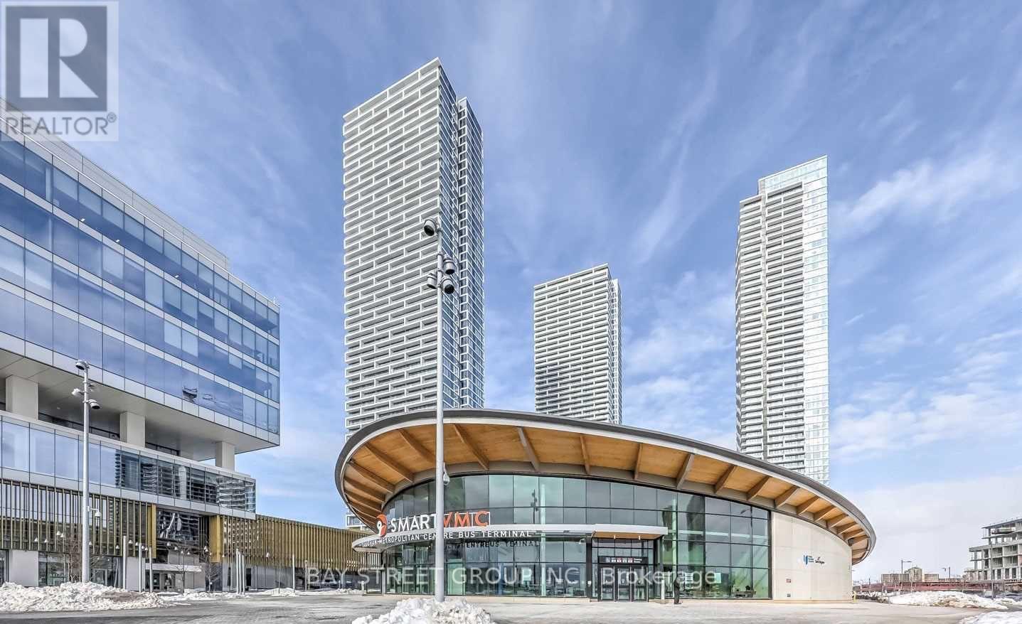 A modern building with a curved glass entrance and three tall towers in the background.