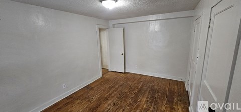 A room with wooden floors and white walls, with a door on the left and a wall-mounted light fixture.