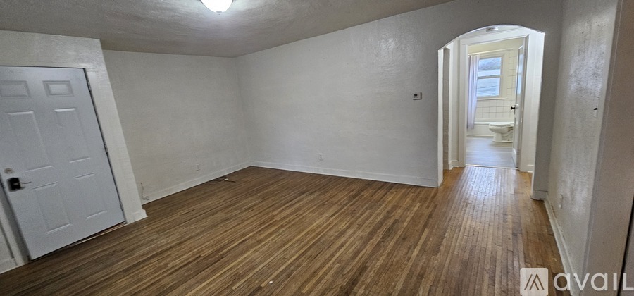 A room with wooden floors and white walls, with a door on the left and an archway leading to another room on the right.