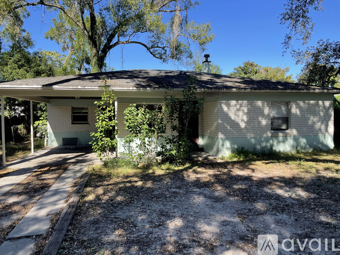 A house with a porch and a covered patio is surrounded by trees and has a gravel driveway.