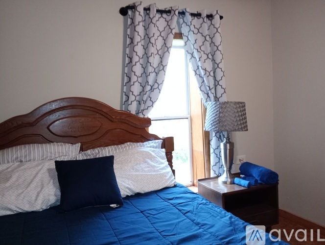 A bed with a blue comforter and pillows is in front of a window with curtains.