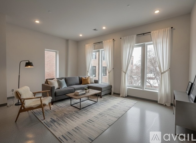 A living room with a grey couch, a wooden chair, and a large window with white curtains.