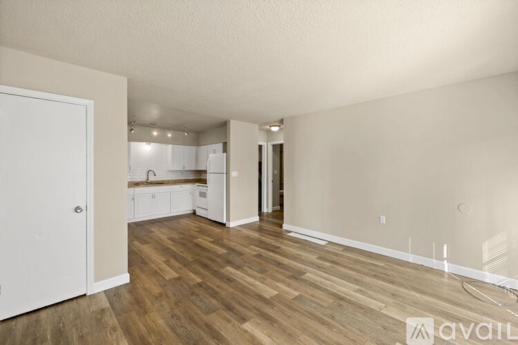 A room with wooden floors and white walls, with a kitchen visible in the background.