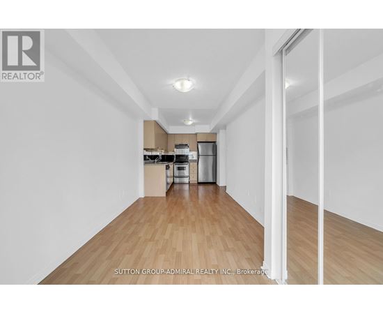 A spacious room with wooden flooring and white walls, leading to a kitchen area.