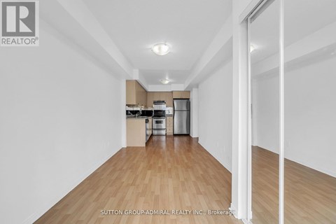 A spacious room with wooden flooring and white walls, leading to a kitchen area.