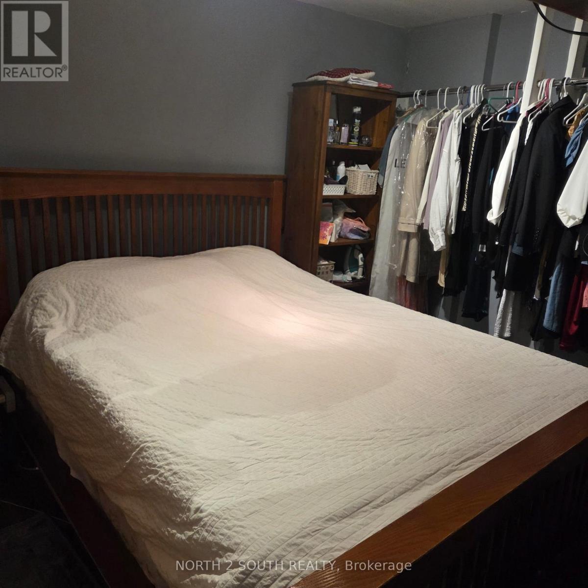 A bed with a white sheet is in the foreground of a room with a closet full of clothes.