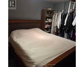 A bed with a white sheet is in the foreground of a room with a closet full of clothes.