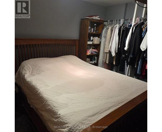 A bed with a white sheet is in the foreground of a room with a closet full of clothes.
