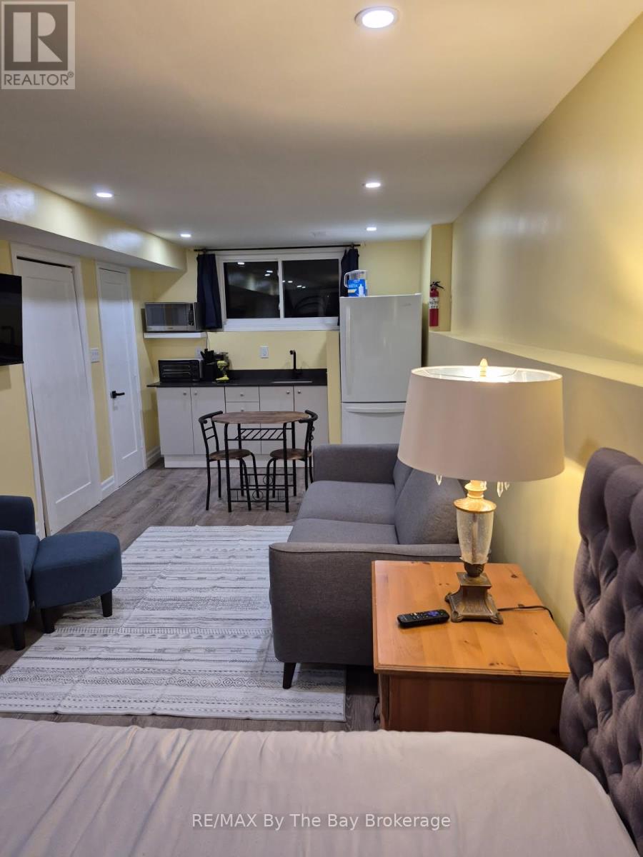 A well-lit hotel room with a bed, a couch, a chair, a table with a lamp, and a kitchenette in the background.
