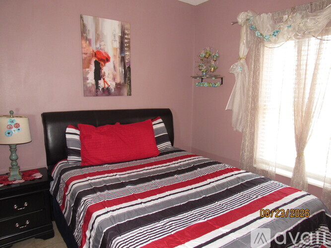 A bedroom with a bed, a nightstand with a lamp, a picture on the wall, and a window with curtains.